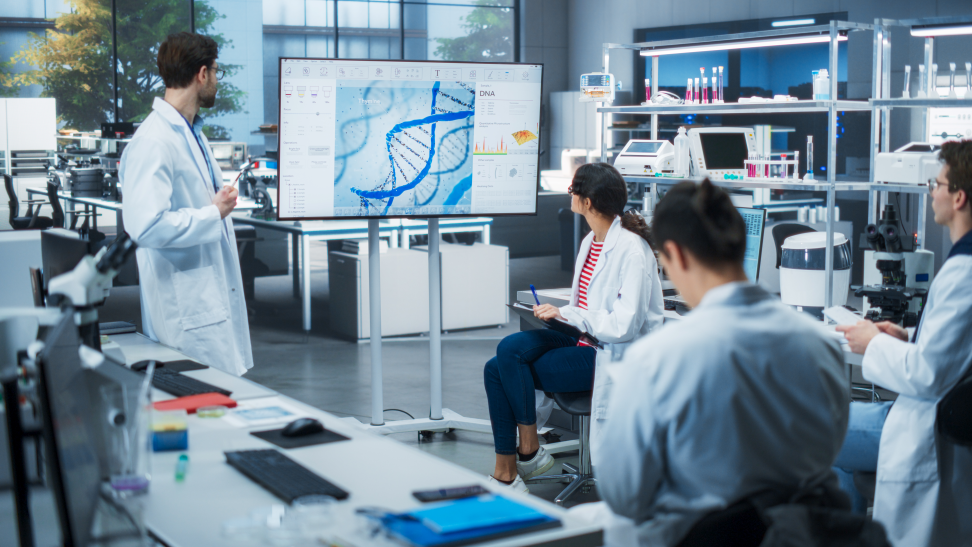A group of individuals in a lab, wearing white lab coats, reviewing data on a large screen.
